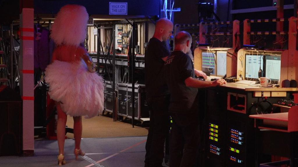 The back of a performer dressed in a white feather head piece carrying feather fans under her arm. Two men dressed in black are looking away from the camera who are working backstage with computer monitors in front of them on a desk.