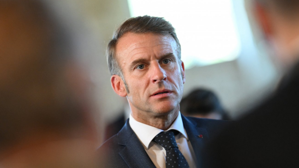 Close up of Emmanuel Macron in dark suit with two people in the foreground, blurred