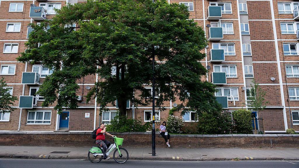 A block of social housing flats viewed from the road, with a man cycling in front of them and a large leafy tree behind him, in front of the flats.
