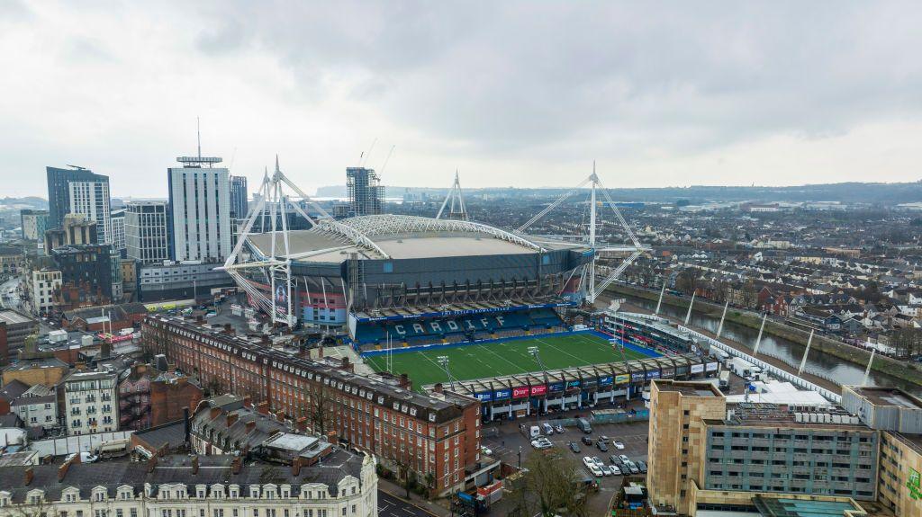 Aerial shot of the Principality stadium in Cardiff. The river taff runs through the city on the right hand side, while the green Cardiff Arms Park football pitch is in front of the stadium. High rise buildings and houses surround the stadium. 