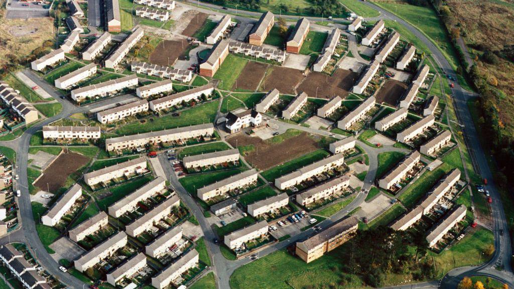 An aerial photo of part of the Gurnos housing estate in Merthyr Tydfil, taken in the 1980s. Rows of white terraced houses are linked by roads, with green space around them. Several patches of mud can be seen where more houses are due to be built.