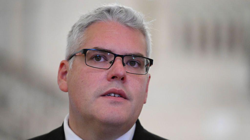 A man with short grey hair wears black glasses with clear lenses 