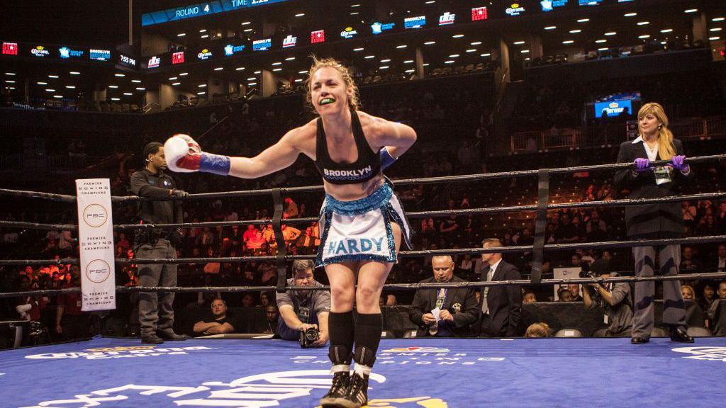 Heather Hardy holds out her arm and bows in a boxing ring