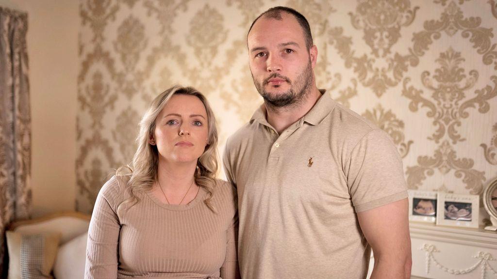 Heidi and Dale standing close together in a living room with cream patterned wallpaper behind them. Pregnancy scan photos are framed on the fireplace. Heidi has long, wavy blonde hair and is wearing a ribbed, long sleeved top and gold necklace. Dale has short dark hair with a beard and is wearing a cream polo-shirt.