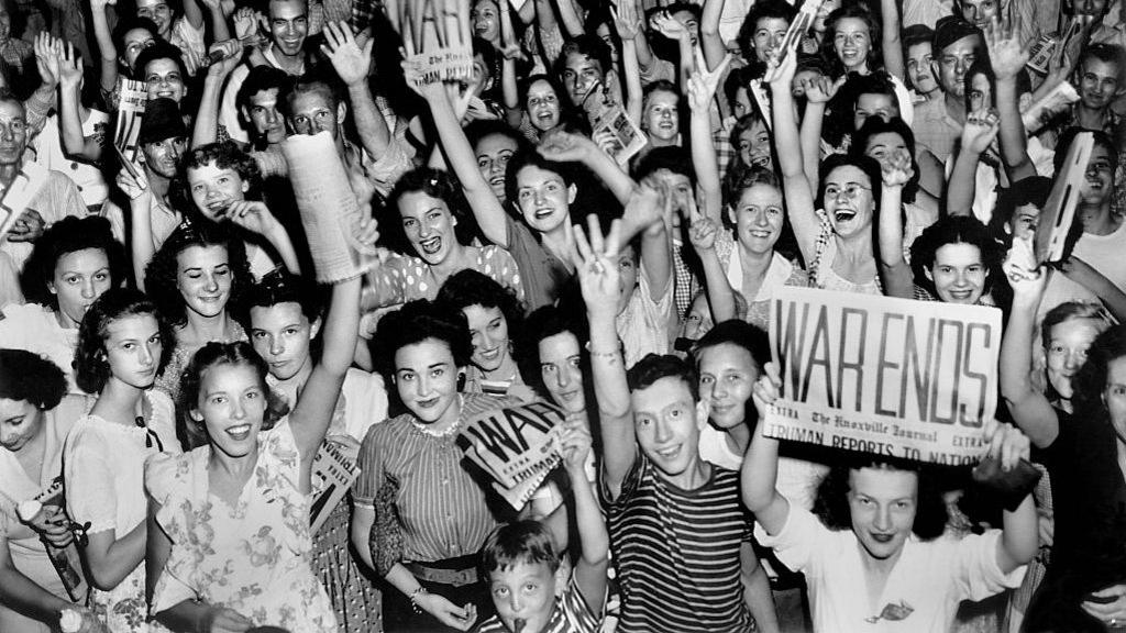Black and white image of large crowd of young women and some men smiling at the camera. One brown-haired woman in the bottom right of the image holds up a newspaper with a large headline reading War Ends