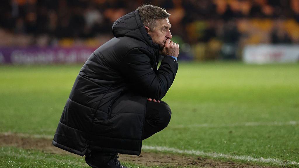 Port Vale manager Jon Brady crouches and looks on from the touchline during a game