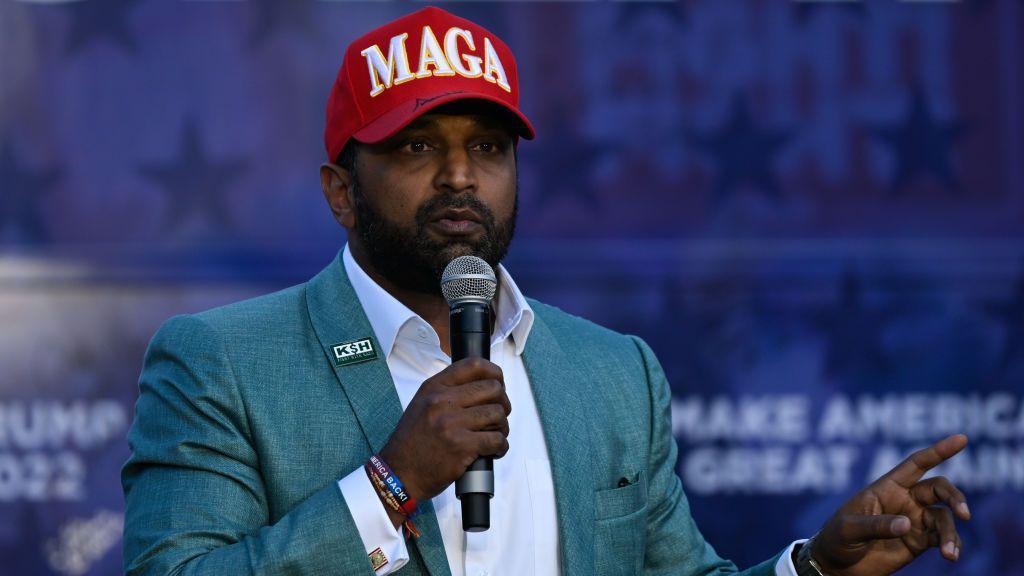 Kash Patel speaking into a microphone wearing a MAGA hat.