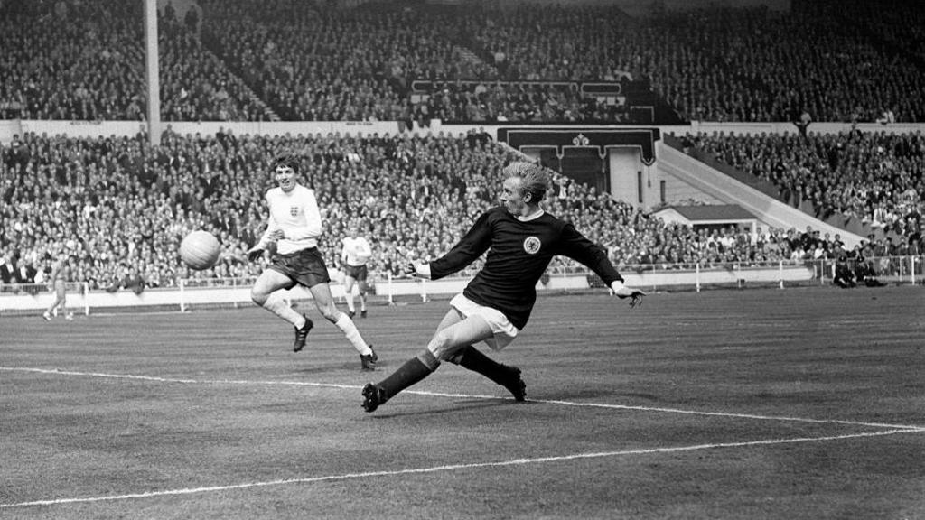 A black and white picture of Denis Law with the ball in a dark Scotland strip. A rival England player stands off to the side and the stands are full of supporters.