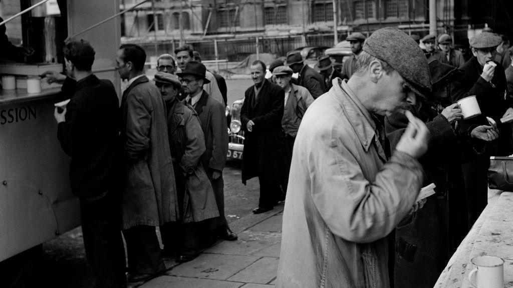 Black and white archive photo of men in long coats eating bread and drinking soup from mugs