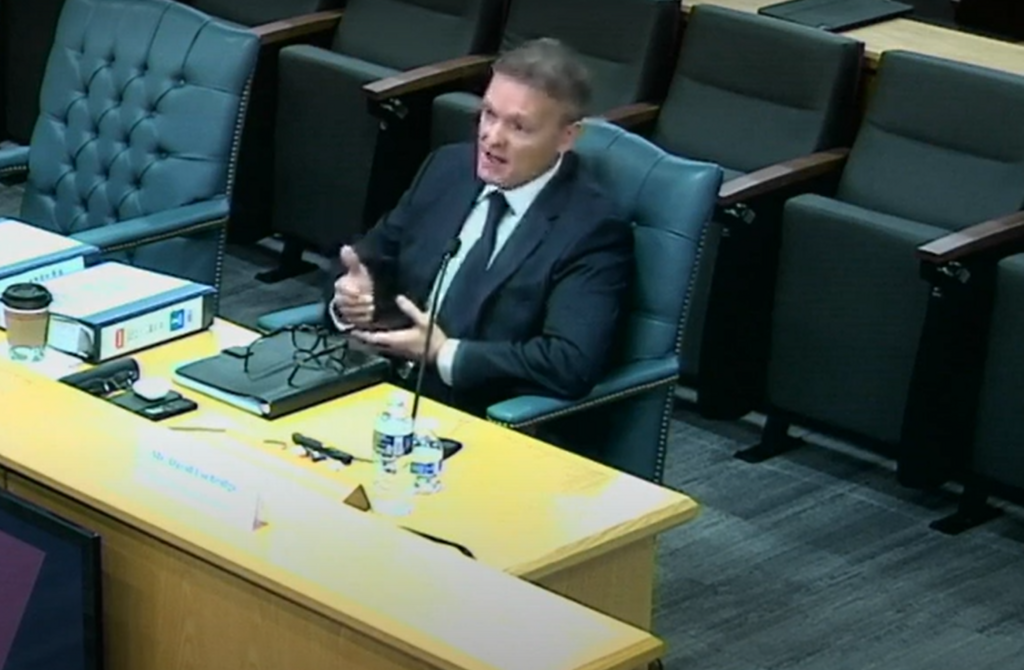 A man in a suit gives evidence to a hearing, surrounded by many empty seats. 