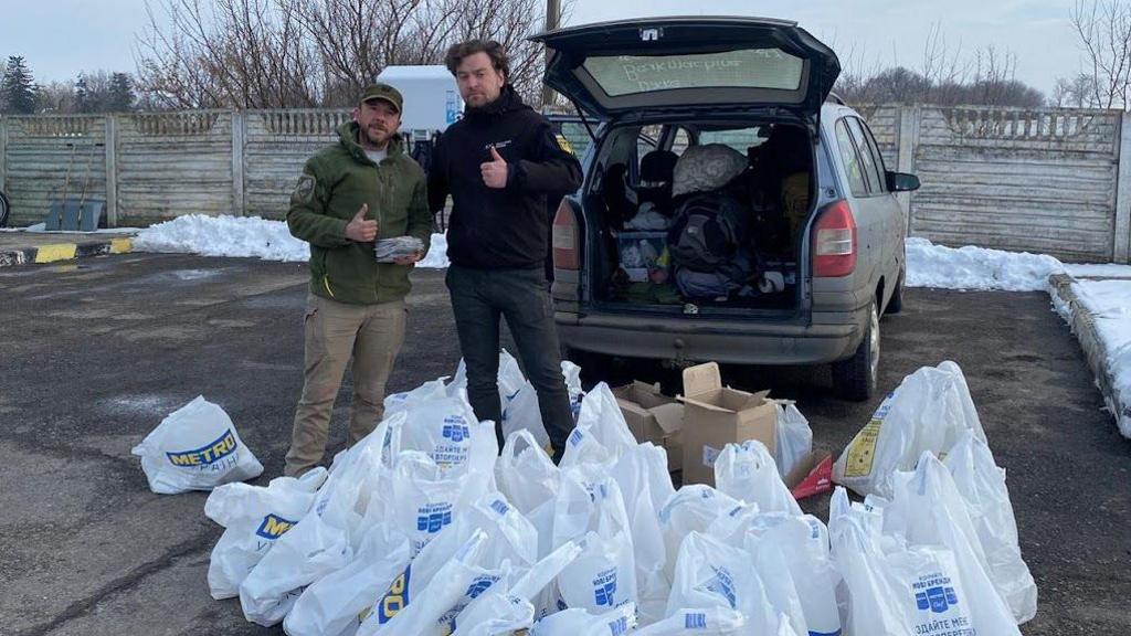 Two men stand behind a large collection of shopping bags on the floor in front of an open car boot. 