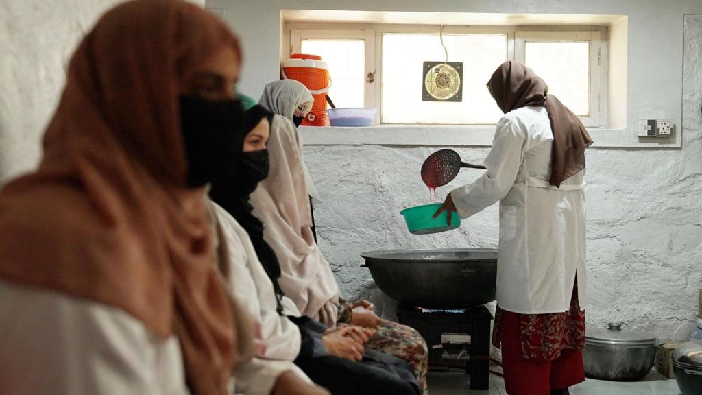 A woman uses a large ladle to transfer jam from a big bowl to a smaller one, as women sit in a line next to her