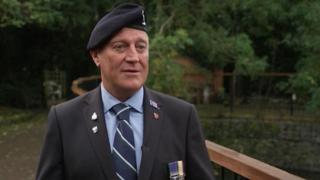 A man wearing a beret and a dark suit with a blue shirt and navy striped tie. His jacket is adorned with medals and pin badges. He is standing next to a wooden railing and there are trees in the background