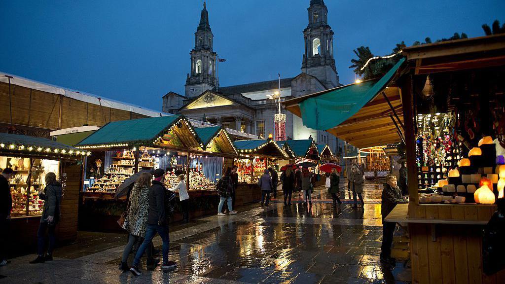 Leeds Christmas market at night, in the rain, showing people walking past wooden cabins selling Christmas-themed gifts with Leeds Civic Hall in the background