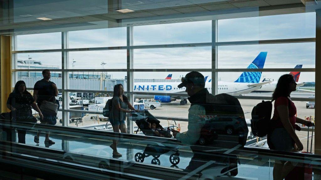 People walk through Ronald Reagan airport in Washington. A woman is pushing a pram and others can be seen in silhouette. out the window are planes waiting on the tarmac.