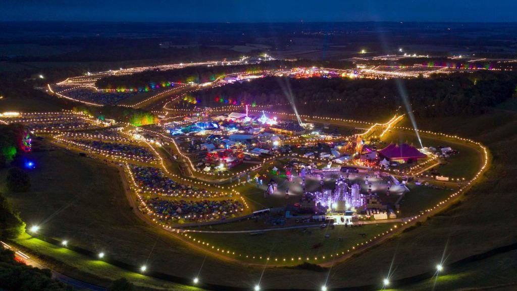 An aerial photo of a festival site and night. There are lots of lit-up tents and everything is enclosed in a fence of fairy lights.
