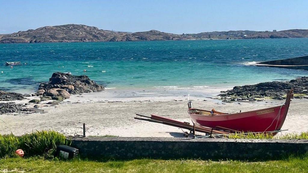A turquoise sea meets a sandy beach which has a red boat parked on it. There are rocky hills in the background.