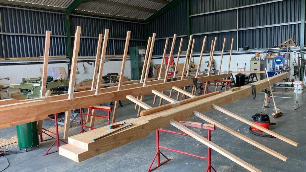 Wooden sail frames can be seen propped on red metal stilts within a workshop. The frames have prongs of wood that spread horizontally off one main wooden plank.