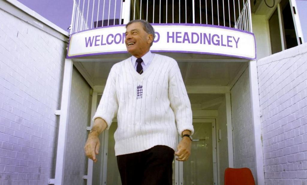 Man with grey hair wearing a white knitted cricket jumper walks under a sign surrounded by stadium. A sign above declares 'Welcome to Headingley'.