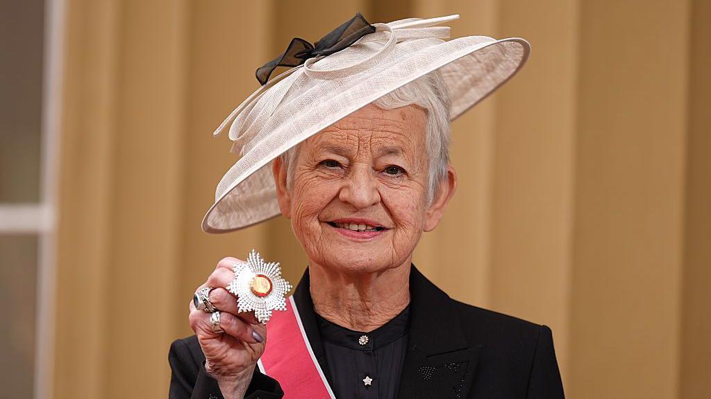 Jacqueline wilson posing with her grand cross medal wearing a white wide rimmed hat and black suit
