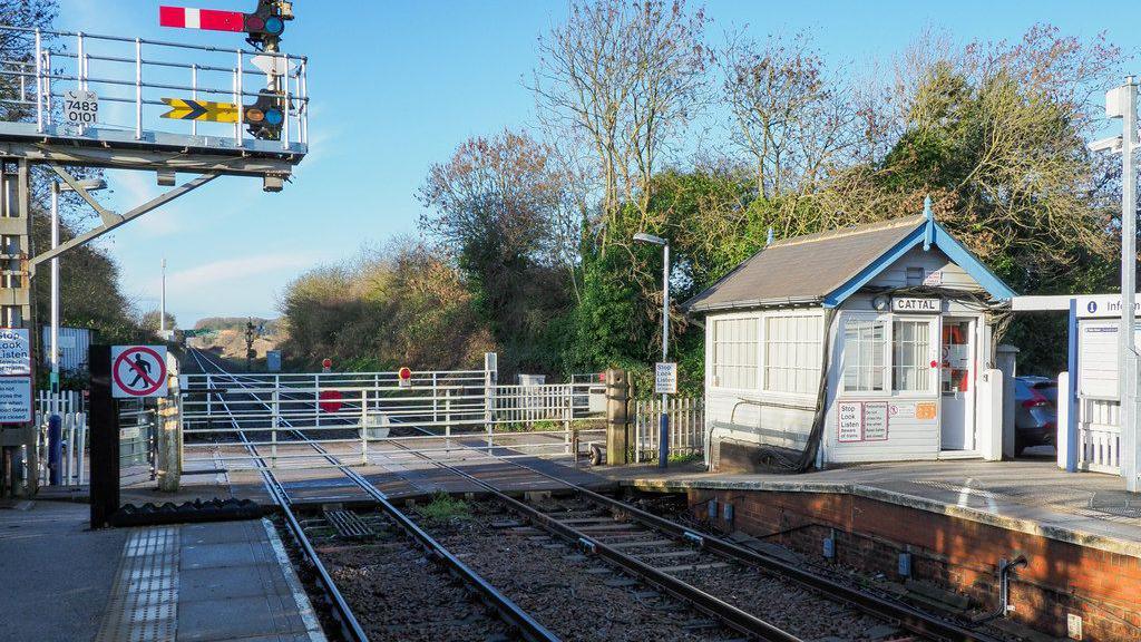 Cattal railway station. The picture shows a level crossing at the end of the platform. There are two signals in the picture as well as a signal box. 