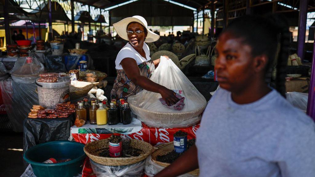 A woman at a market stall with a wide-brimmed hat on cleans a clear plastic bag protecting a basket of sugar. 