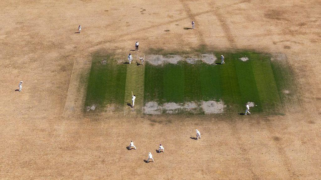 A cricket match on a yellow-brown, dry outfield with a green square