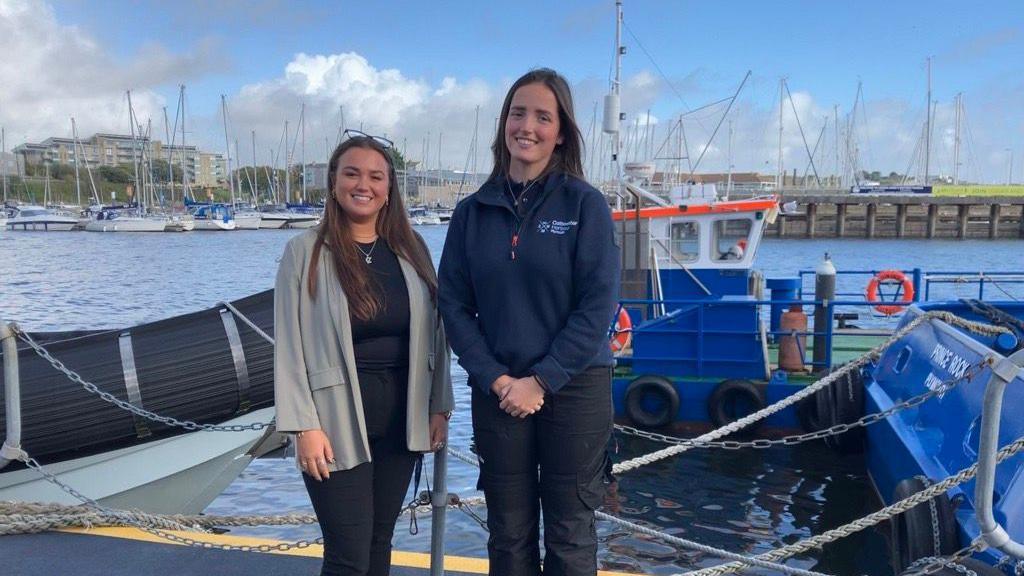 Evie Smith on the left and Emma Maunder on the right stood together in front of the sea with a blue fishing boat behind. Evie is wearing a black top and black trousers with a green blazer, and Evie is wearing a navy blue jumper with black trousers. They are both looking at the camera and smiling.