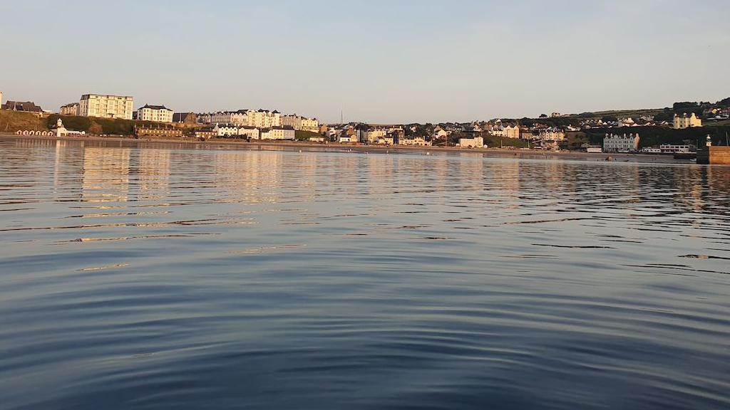 Sea swimmers in the distance and Port Erin beach in the background, with calm waters and a light coloured sky.