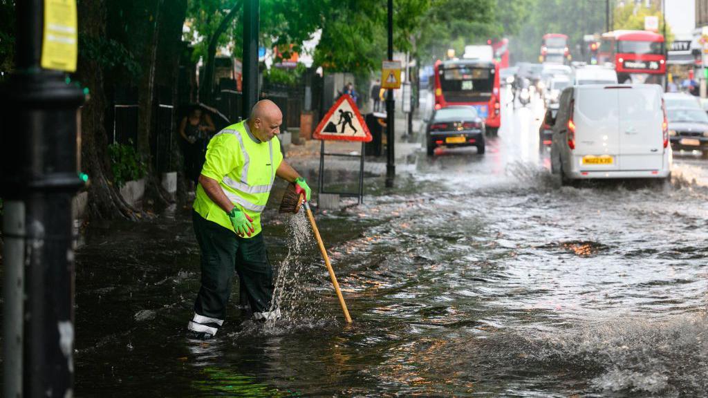 A council worker in a high viz jackets tries to tackle a flooded road in London. He is using his broom to try to clear a drain. Cars and buses are driving past through the flood waters.