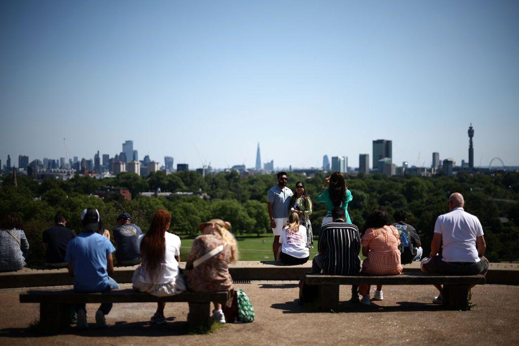 People are sat on benches looking at the view of London's skyline from Primrose Hill Park. In the background a man and woman are having a photo taken together.