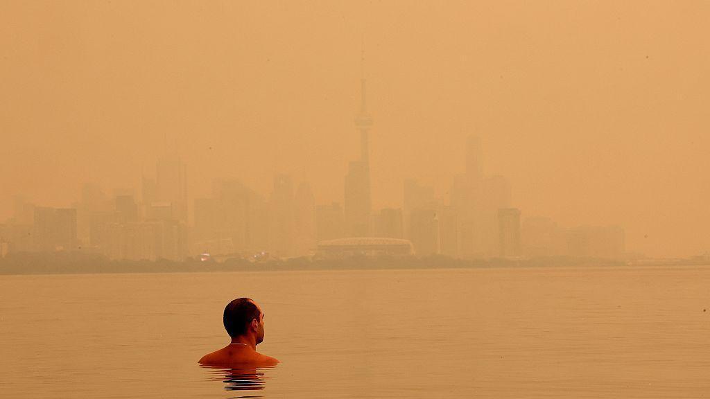 A man swimming in Lake Ontario on 15 July overlooking the Toronto skyline blanketed by a bright, orange haze from wildfire smoke. 