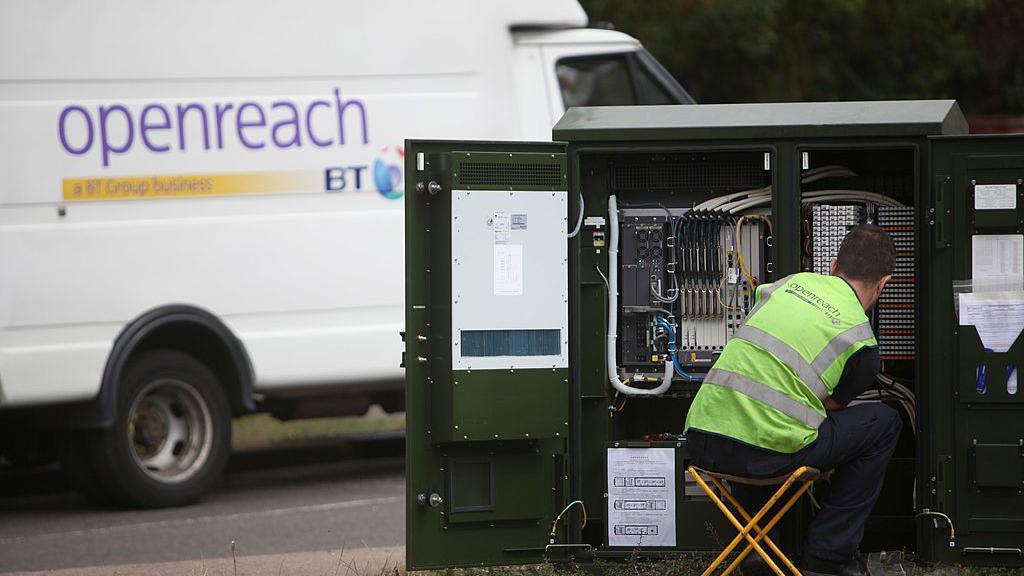 An Openreach employee with a hi-vis jacket on working on cables of a green broadband street box while a openreach van sits in the foreground. 