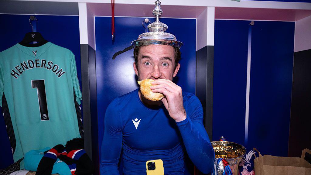 Ben Chilwell celebrates winning the FA Cup with Crystal Palace by putting the trophy lid on his head while eating a burger