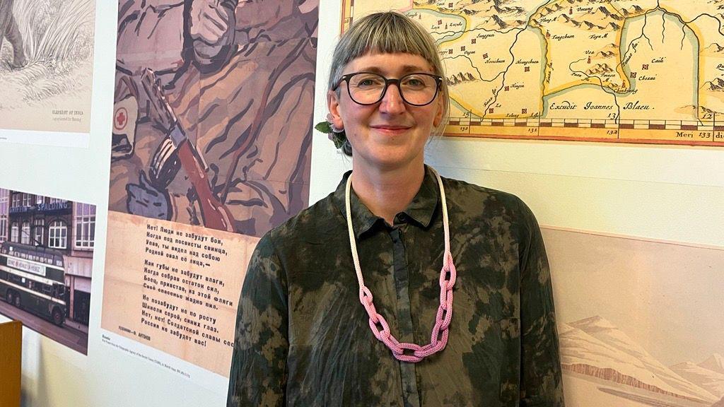 A woman with short grey hair and a short full fringe and black-rimmed glasses is wearing a green and black long-sleeved shirt, chunky pink necklace and smiling into the camera. Behind her is a map and several posters on the wall.