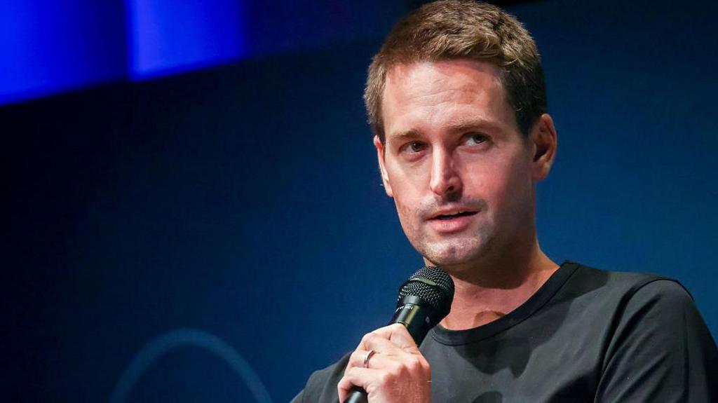 Snap boss Evan Spiegel shown looking away from the camera to his left as he speaks into a microphone on-stage at an event.