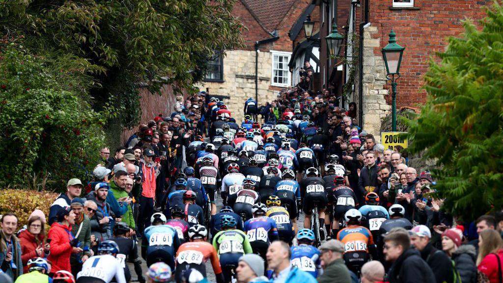 Riders at the Lincoln Grand Prix head up Michaelgate towards the cathedral and Castle Square. Large crowds are standing at each side of the cobbled road with the cyclists in the middle.