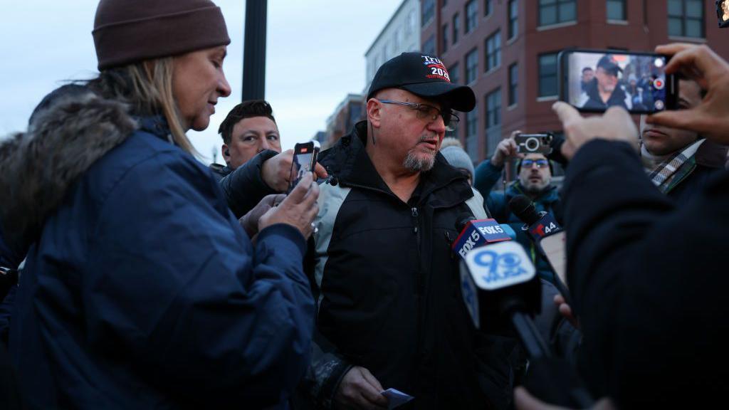 Stewart Rhodes in black shirt and black Trump baseball cap,  surrounded by people holding phones, microphones and cameras