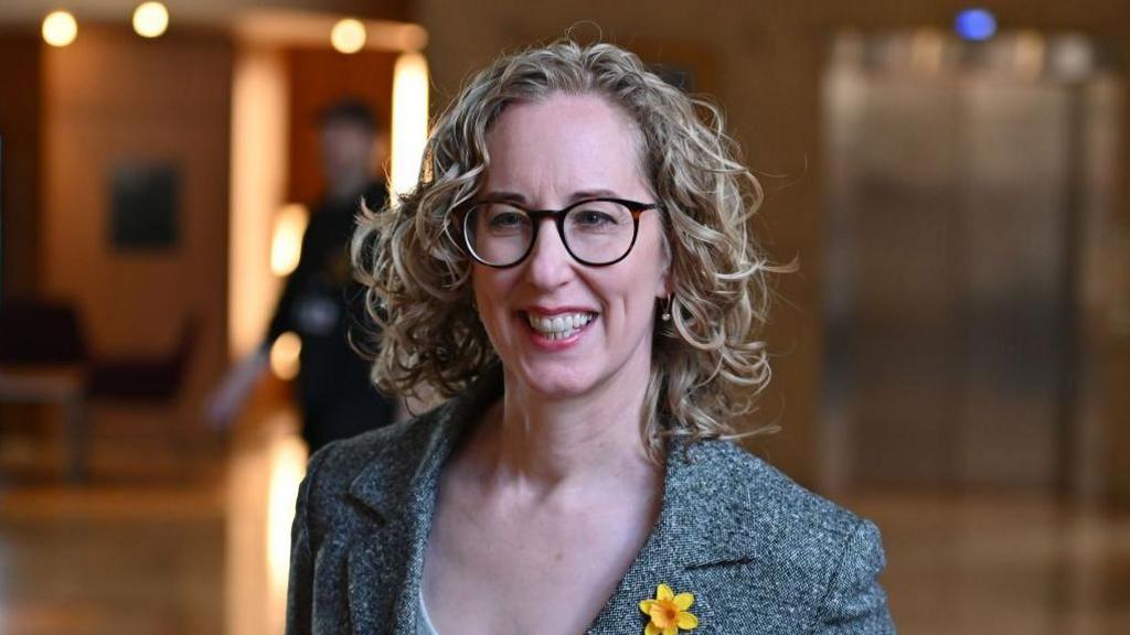 Lorna Slater - a woman with curly blonde hair and glasses, wearing a grey suit jacket with a yellow flower on it - pictured at the Scottish Parliament
