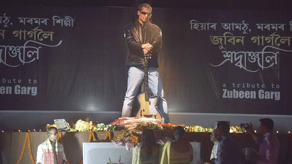 A huge poster of Zubeen Garg - short hair, sunglasses, black jacket, blue jeans and holding a guitar. fans gather before the poster to pay tribute to him.