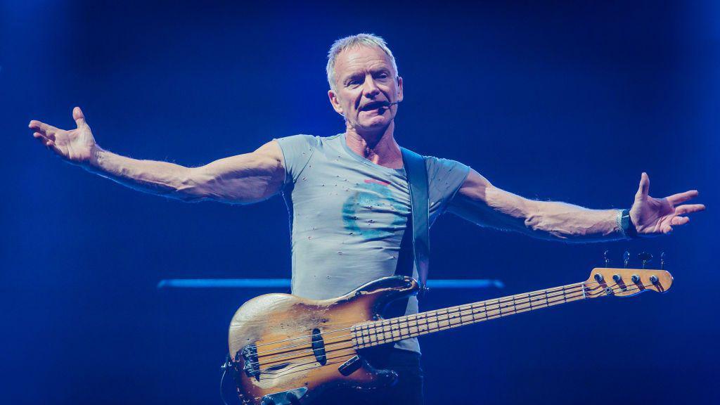 Sting is pictured performing on stage during a show in Milan, Italy. He has his arms stretched out wide while he sings and a guitar hangs from his body.