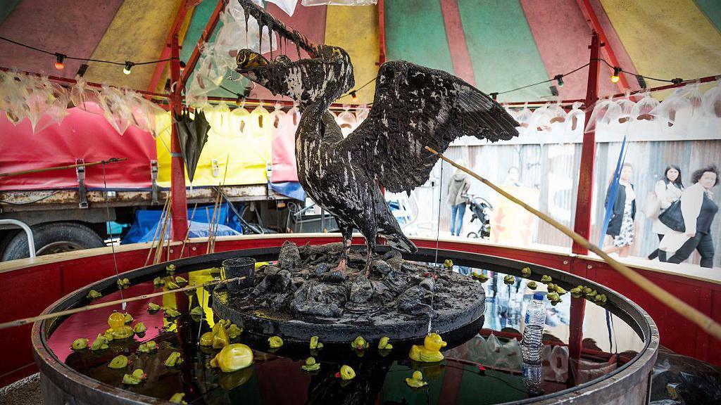 The traditional hook-a-duck game at fairgrounds, but with  twist. Below a colourful striped canvas tent, there is a large white seabird perched on rocks in the centre of a vat of dark oil, which it is partially covered in. Its wings are outstretched and its beak is open, with crushed drink cans crammed inside its mouth. Below it are small rubber ducks which have overturned in the grease. It is likely a commentary on the damage oil spills can inflict on ecosystems and wildlife. 