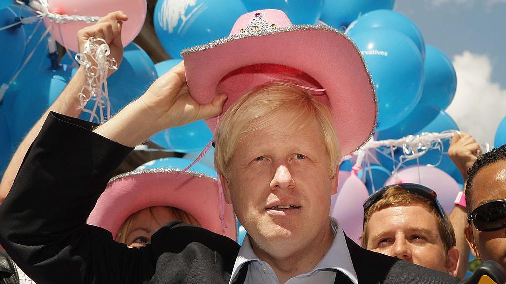 Boris Johnson holds a pink Stetson hat above his head. He is wearing a blue shirt and a dark suit jacket. 