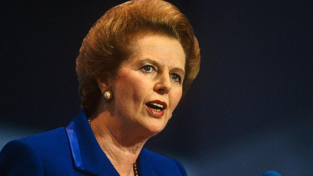 Margaret Thatcher is pictured with a steely expression staring forward as she talks to an audience. She has light red hair, back-combed and rounded into a bob and she is wearing a blue jacket.