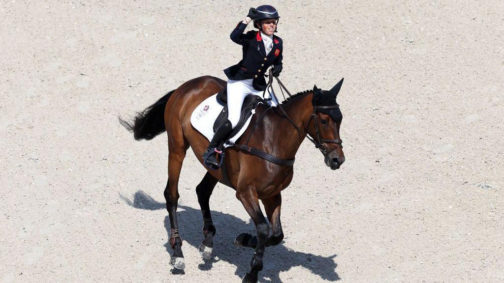 A woman in a formal horse riding outfit punches the air as she canters around the sandy arena on a dark brown horse. It is a sunny day and the horse and rider’s shadow is clear. 