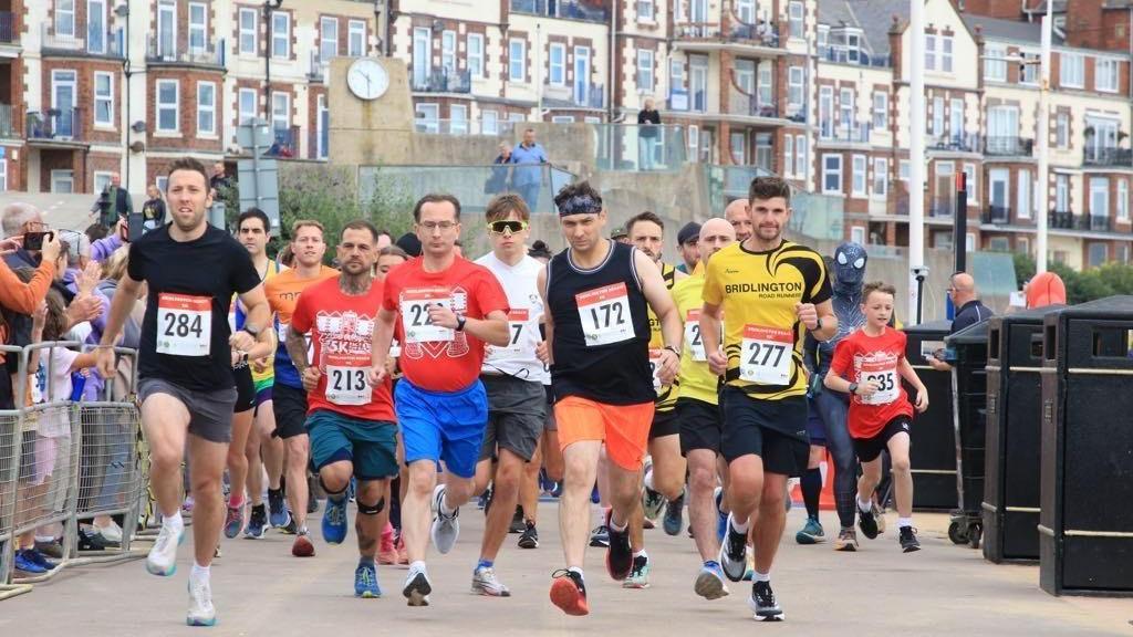 Male runners with numbers pinned to their tops head towards the photographer along a seaside promenade, with apartment buildings in the background. Crowds stand behind barriers at the sides. One runner wears a grey Spiderman outfit.