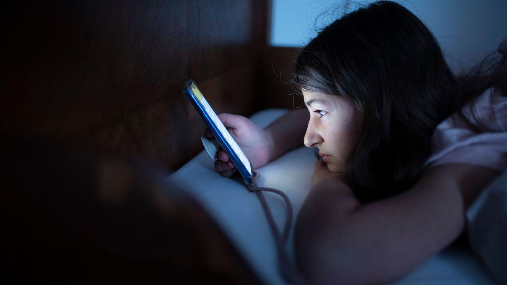 A teenage girl lying on a bed using a smartphone in a dark room