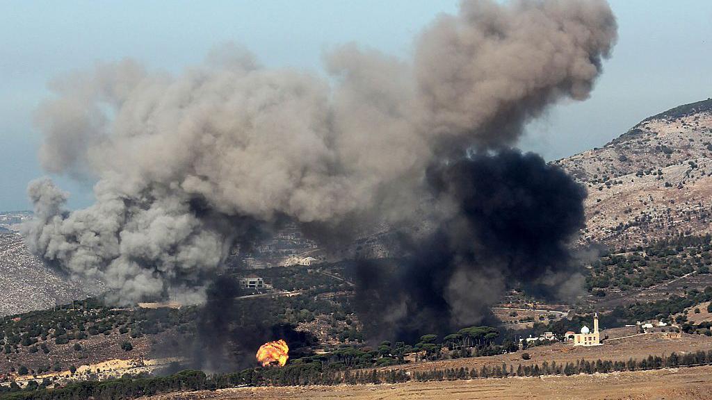 Smoke rises over a wooded area on an arid slope in daytime.