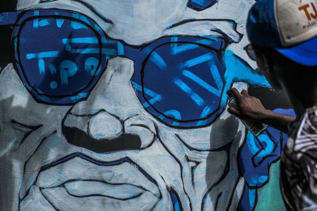 An artist spray-paints a large mural of a man wearing blue sunglasses.