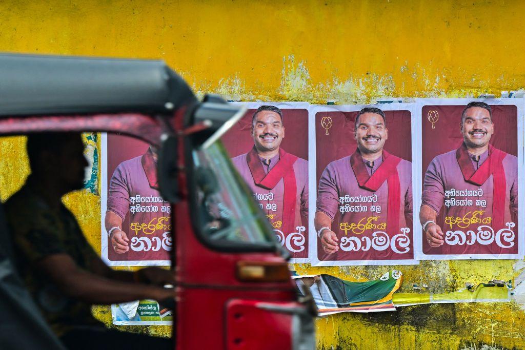 A red motorcycle taxi drives past a yellow wall with posters of a man in a red scarf